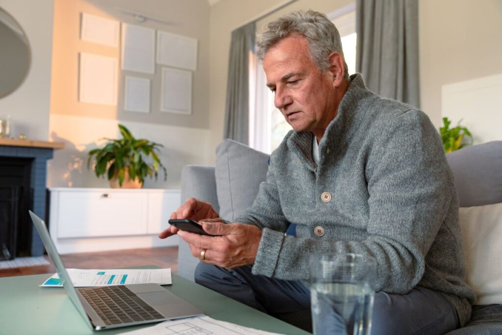 Un homme âgé utilise un smartphone et un ordinateur portable pour lire sur l'augmentation des cotisations à la RRQ en étant assis sur le sofa.