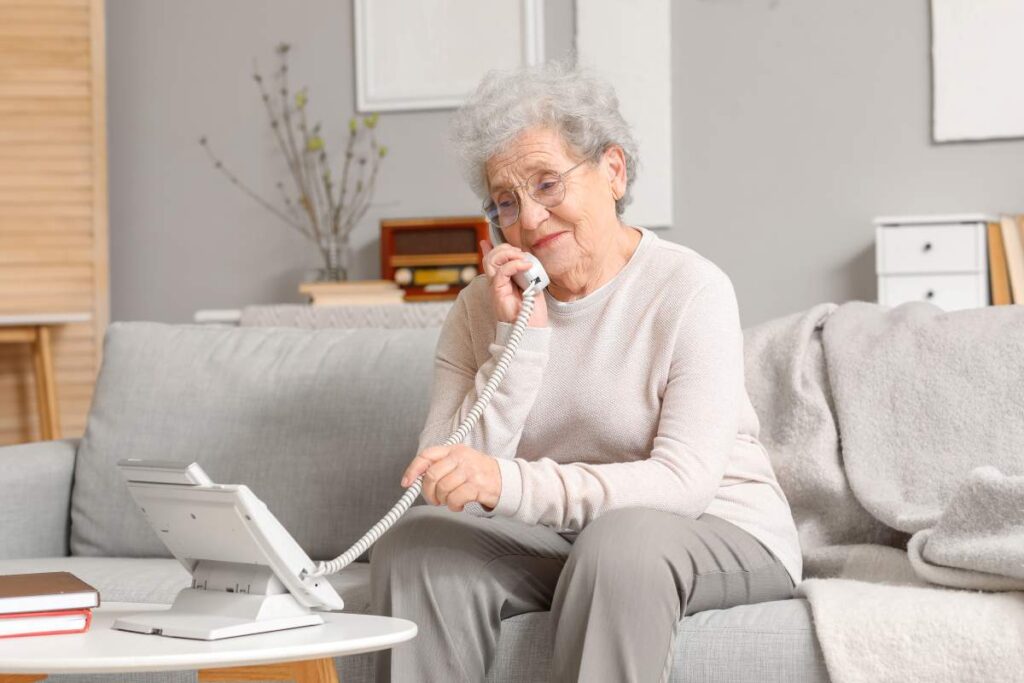 Une femme âgée est assise sur un canapé et parle au téléphone, illustrant une démarche simple pour contacter Retraite Québec depuis chez soi.